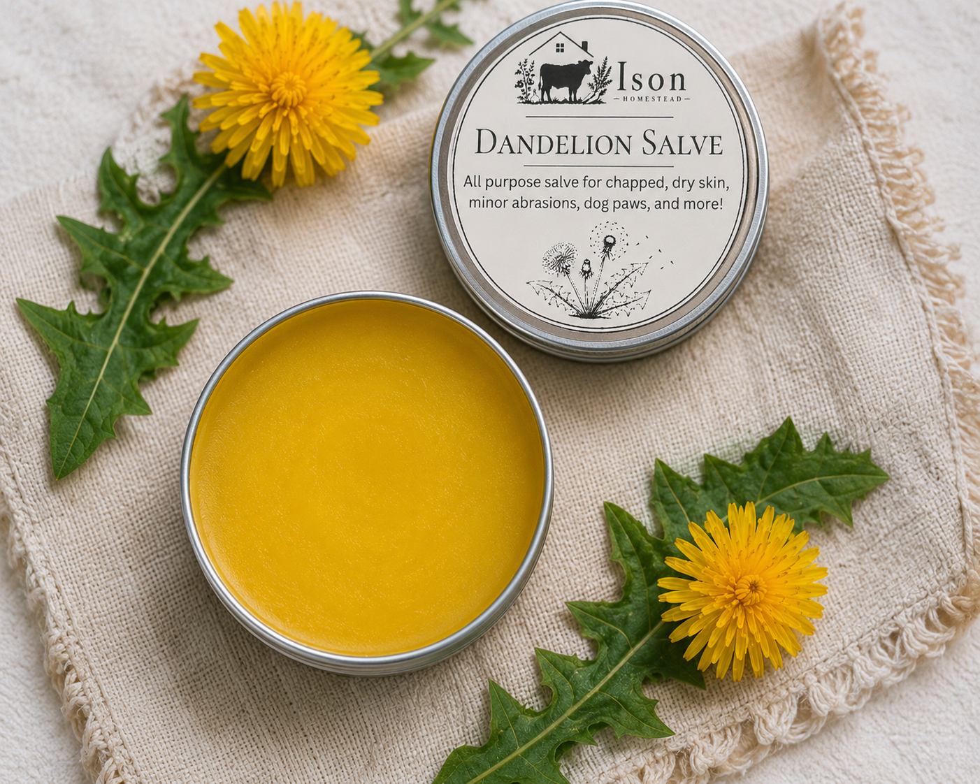 Dandelion salve in a metal tin with dandelion flowers and leaves on a textured fabric background.
