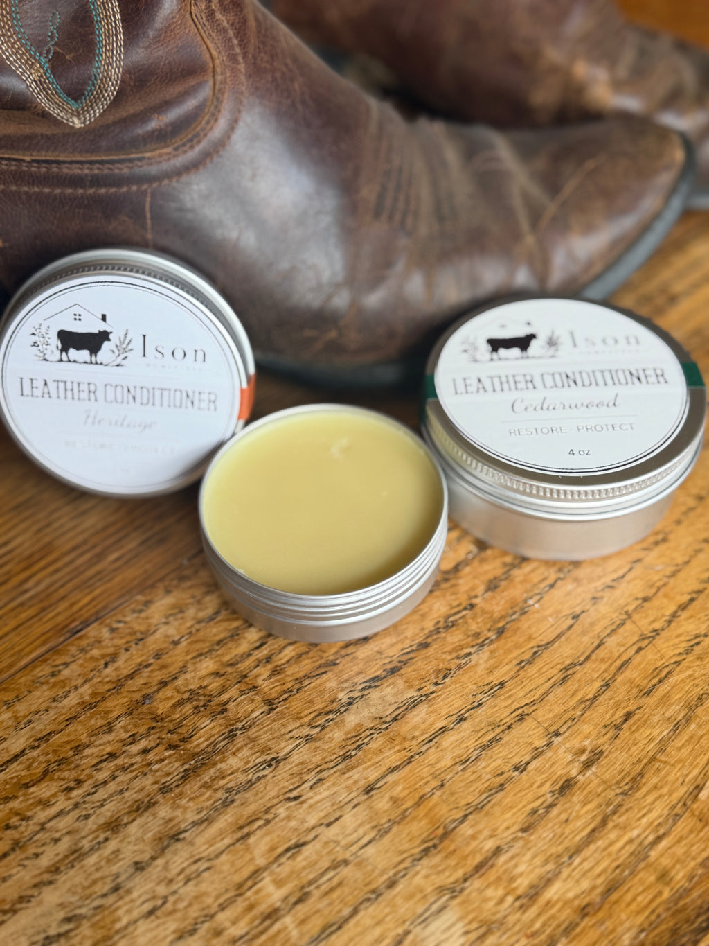 Two tins of leather conditioner with a boot in the background on a wooden surface.