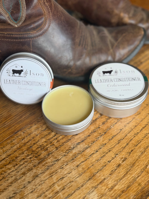 Two tins of leather conditioner with a boot in the background on a wooden surface.